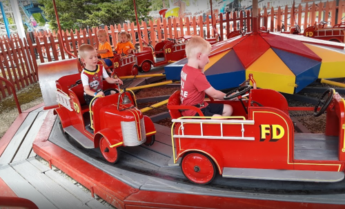 Two young children ride on red fire truck-themed amusement rides in a colorful, fenced play area.
