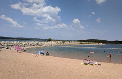 A sandy beach by a calm lake, with people swimming and relaxing under a blue sky with scattered clouds.