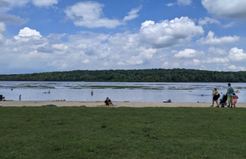 A sandy beach by a calm lake, with people swimming and enjoying the sunny day under a partly cloudy sky.