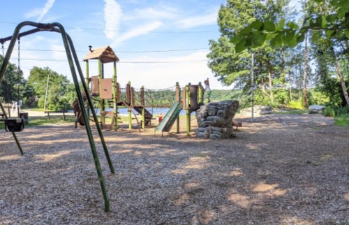 A playground with swings, a climbing structure, and a slide, surrounded by trees and a gravel surface.