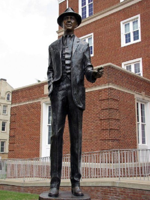 A bronze statue of a man in a suit and hat, standing confidently in front of a brick building.