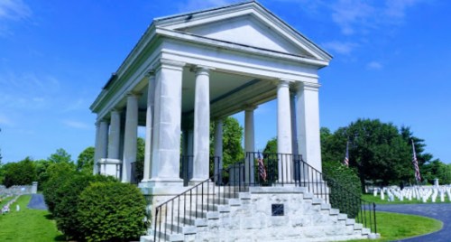 A white marble gazebo with columns, surrounded by greenery and gravestones, under a clear blue sky.