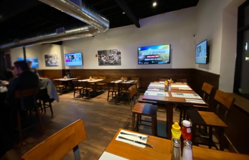 Interior of a restaurant with wooden tables, chairs, and multiple screens displaying content on the walls.