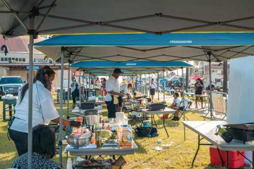 A bustling outdoor cooking event with chefs under tents, preparing food and interacting with attendees.