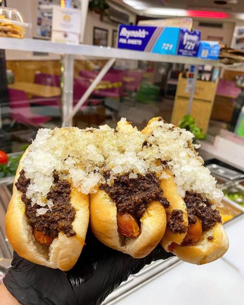 Three hot dogs topped with a generous layer of chopped onions and a rich, dark chili sauce, held in a gloved hand.