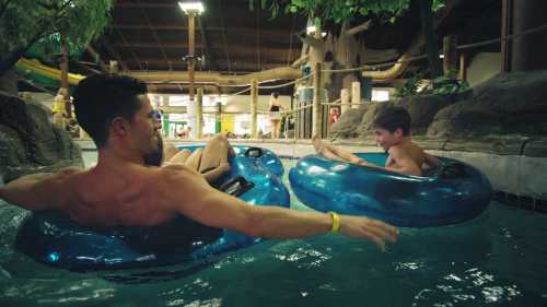 A man and a boy float on blue inner tubes in a water park pool, surrounded by trees and playful water features.