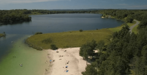 Aerial view of a sandy beach by a calm lake, surrounded by green trees and a clear blue sky.