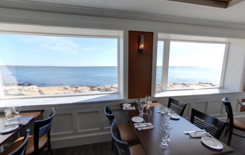 Interior of a restaurant with tables set for dining, featuring large windows overlooking a calm ocean view.