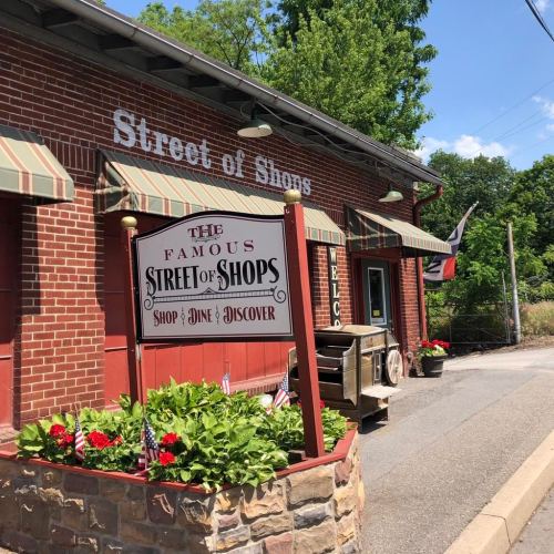 A brick building with a sign reading "Street of Shops," surrounded by flowers and flags, inviting visitors to explore.