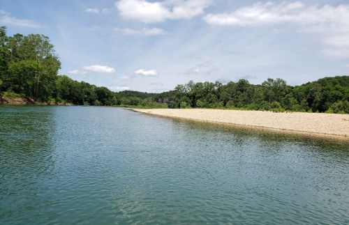 A serene river scene with a sandy shore, surrounded by lush green trees under a partly cloudy sky.