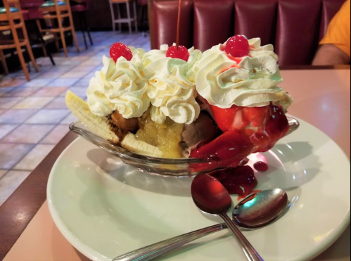 A banana split topped with whipped cream and cherries, served in a glass dish on a white plate. Two spoons beside it.