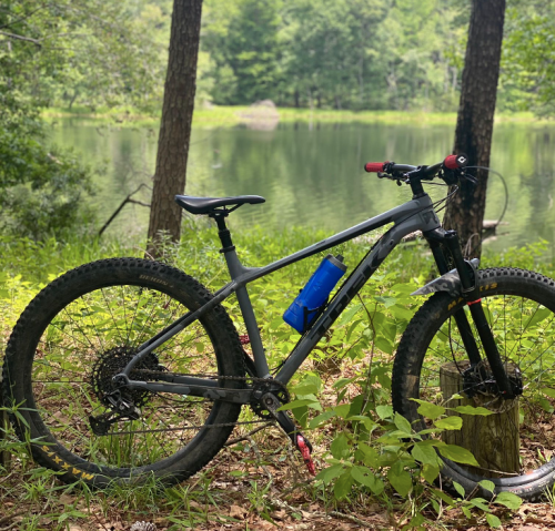 A mountain bike rests on the ground near a serene pond, surrounded by lush greenery and trees.