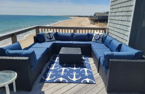 A cozy outdoor seating area with blue cushions, a coffee table, and a nautical rug, overlooking a sandy beach and ocean.