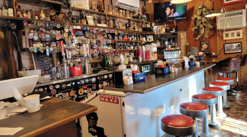 A cozy bar with a wooden counter, red stools, and a wall adorned with stickers and memorabilia.