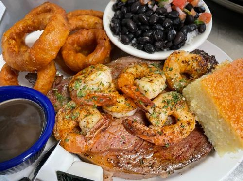 A plate featuring grilled shrimp, steak, onion rings, black beans with pico de gallo, and cornbread.