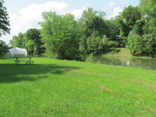 A serene riverside scene with green grass, trees, and a picnic table near a calm waterway under a blue sky.
