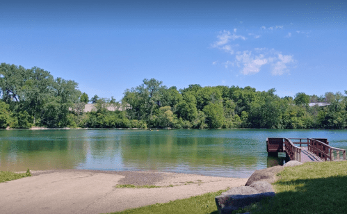A serene lake surrounded by lush greenery, with a wooden dock extending into the water under a clear blue sky.