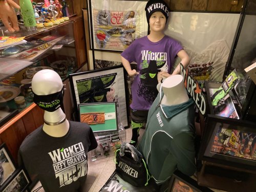 Display of "Wicked" merchandise, including shirts, hats, and posters, in a cozy shop setting.