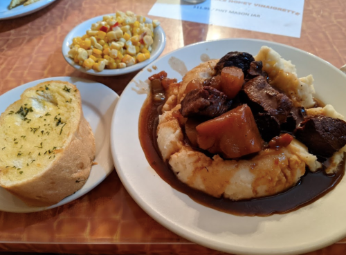 A plate of mashed potatoes topped with beef stew and vegetables, served with garlic bread and a side of corn salad.