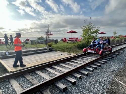 A person in an orange shirt stands by a wooden platform as a group rides a red pedal cart along a railway track.