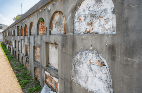 Weathered brick wall with arched openings, showing signs of decay and overgrown grass beside it.
