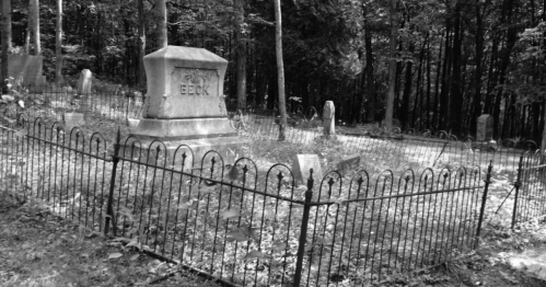 A black and white image of a cemetery with a fenced grave marked "Beck" surrounded by trees and overgrown grass.
