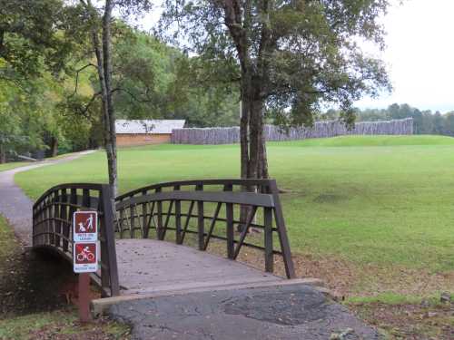 A wooden bridge leads over a path in a park, with a grassy area and a wooden structure in the background.