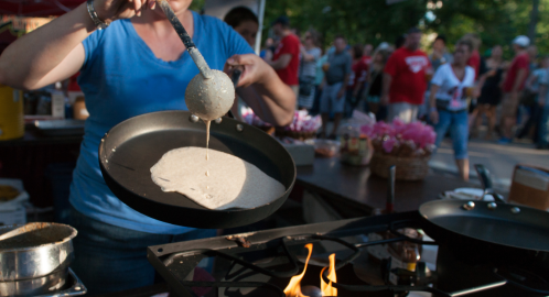 A person in a blue shirt pours batter onto a hot pan at a busy outdoor food festival. Flames are visible beneath the pan.
