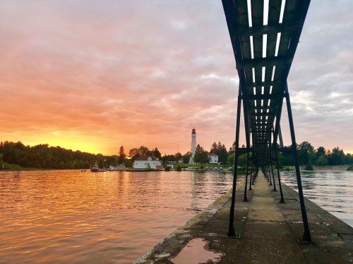 A pier extends over calm water at sunset, with a lighthouse and trees in the background under a colorful sky.