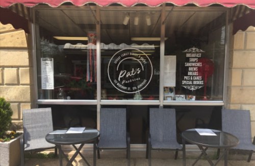 A cozy café window with a sign reading "Pat's Pastries," featuring outdoor seating and a menu displayed.