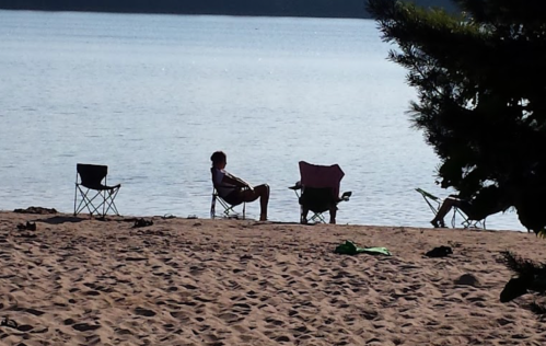 A serene beach scene with several empty chairs and a person relaxing by the water's edge.