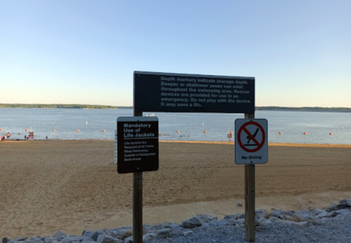 Signage at a beach warns about depth markers, life jacket use, and prohibits driving on the sand. Calm water in the background.