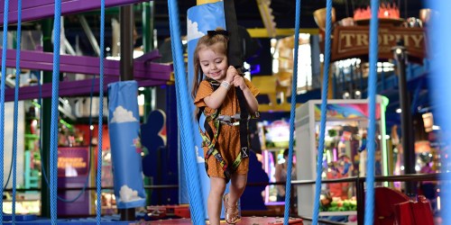 A young girl in a harness smiles while navigating a colorful indoor obstacle course.