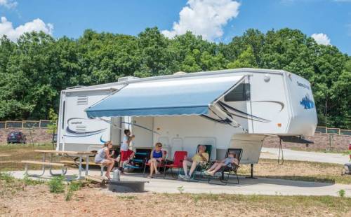 A group of people relaxes outside a parked RV with an awning, surrounded by trees and a picnic table.