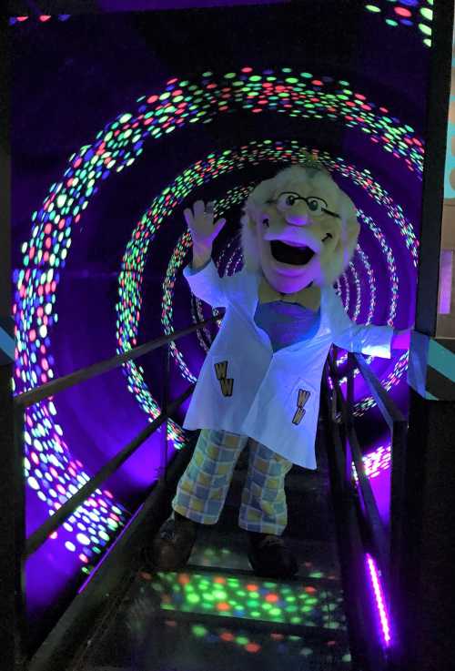 A colorful tunnel with a puppet in a lab coat and glasses, waving cheerfully amidst swirling lights.