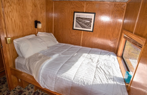 Cozy wooden bedroom with a double bed, white bedding, and a framed photo on the wall. Natural light from a window.