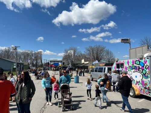 A lively outdoor event with food trucks, families, and children enjoying a sunny day under a blue sky with clouds.