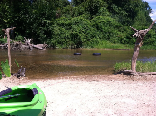 A green kayak on a sandy riverbank with tires floating in the water and lush greenery in the background.