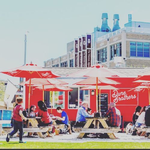 Outdoor dining scene with red umbrellas, people at picnic tables, and a food truck in a vibrant urban setting.