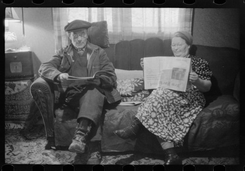A man and woman sit on a couch, reading newspapers in a cozy, vintage living room.