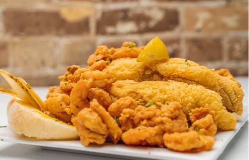 A plate of golden fried fish fillets and shrimp, garnished with lemon wedges and served with a slice of bread.