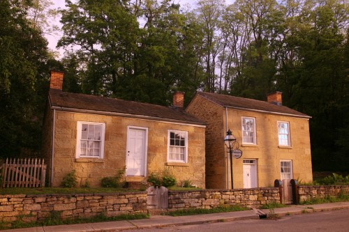 Two historic stone houses with white doors, surrounded by greenery, set against a backdrop of trees at sunset.