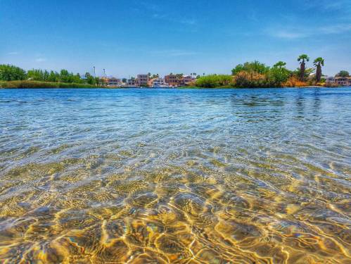 Clear water with ripples in the foreground, lush greenery and buildings in the background under a blue sky.