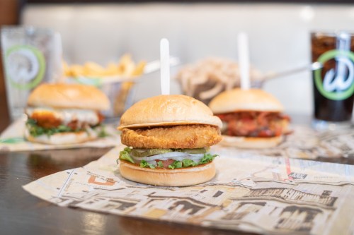A close-up of a crispy chicken sandwich on a bun, with lettuce and tomato, surrounded by drinks and fries in the background.