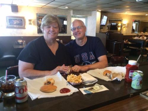 A couple sits at a diner table with burgers, fries, drinks, and condiments, smiling at the camera.