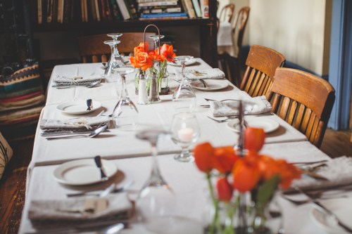 A beautifully set dining table with orange roses, plates, and glassware, surrounded by wooden chairs.