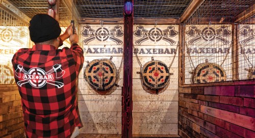 A person in a plaid shirt throws an axe at targets in an axe-throwing venue decorated with wooden walls.