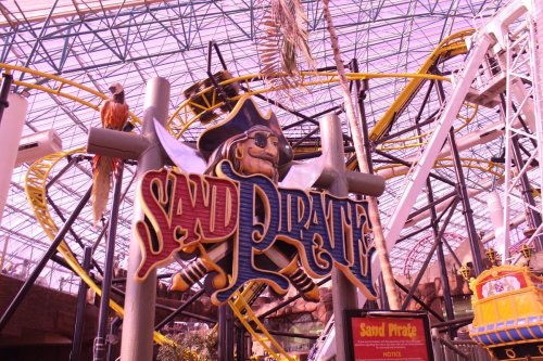 Colorful sign for "Sand Pirate" ride inside a large indoor amusement park with roller coasters in the background.