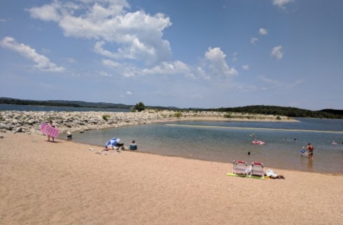 A sandy beach by a calm lake, with people swimming and relaxing under a blue sky with scattered clouds.