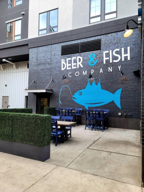 Outdoor seating area with a mural of a fish and the text "Beer & Fish Company" on a brick wall.